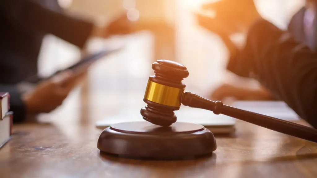 Judge's gavel resting on a wooden desk in a courtroom with blurred figures in the background.