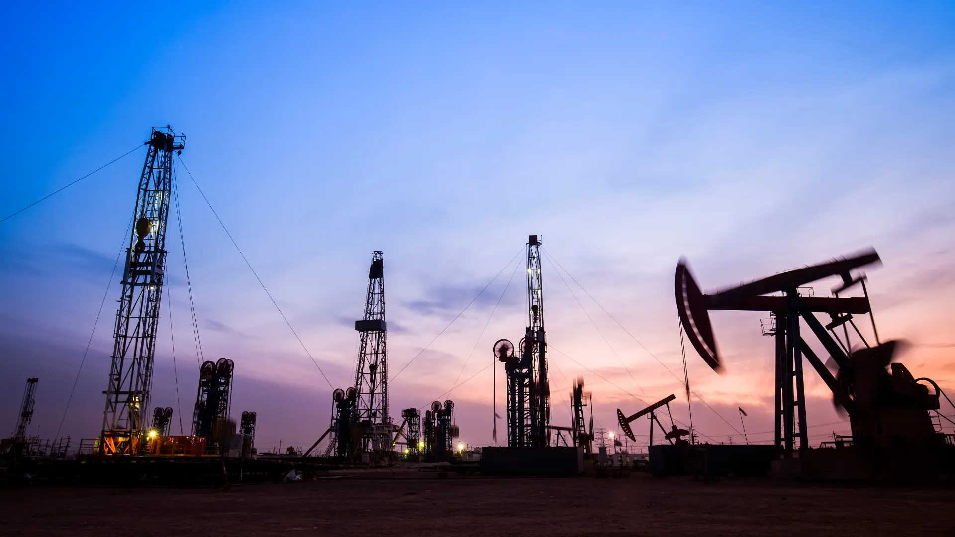 Oil drilling rigs and pump jacks silhouetted against a colorful sunset sky.